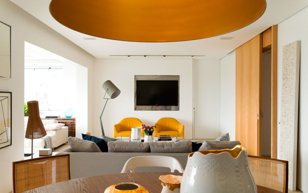 Imagem de sala de estar com cantinho da TV com boas vindas. As poltronas de design, em um escandaloso tom de amarelo brilhante, foram eleitas o ponto de partida para harmonizar os ambientes integrados. O conjunto desse espaço múltiplo – somando os demais elementos, bem como a estrutura de aço inoxidável que envolve o aparelho de TV ao centro –, ganhou mais vibração e jovialidade.  